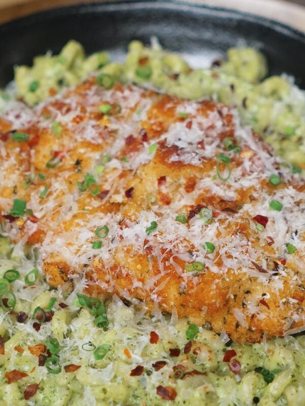 Close-up of golden, crunchy chicken breast resting on a bed of creamy pesto fusilli, garnished with parmesan and fresh green herbs.