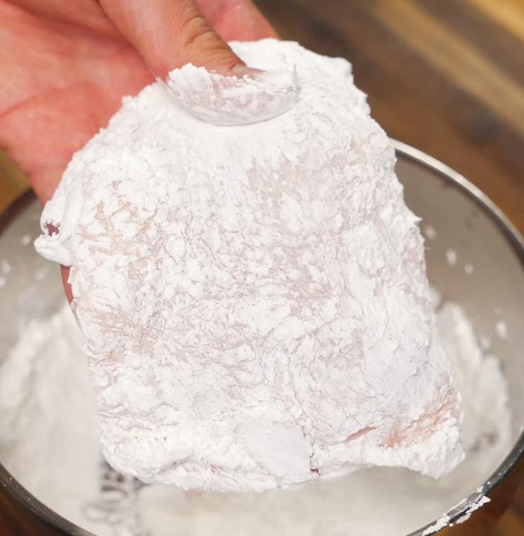 A raw chicken thigh is fully coated in potato starch, ready for frying to achieve an ultra-crispy texture in the honey lemon chicken recipe.