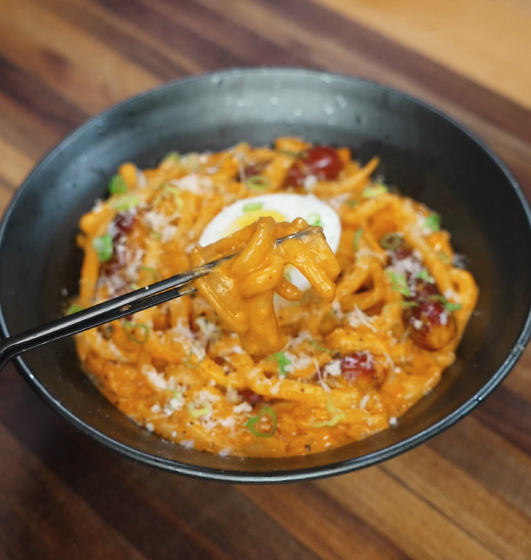 Chopsticks lift a glossy bite of creamy rose noodles from the bowl, highlighting the rich sauce clinging to each strand with sausage and garnishes in the background.