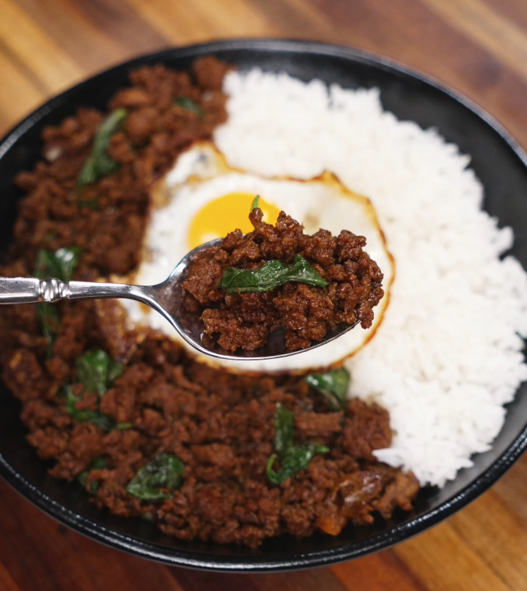 A close-up shot of a spoonful of Thai pad kra pao with bits of crispy basil and ground beef lifted over a bed of jasmine rice and egg.