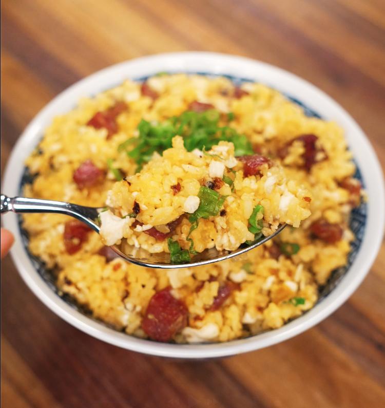 A macro image of a spoonful of golden fried rice being lifted from a full bowl, showcasing fluffy rice grains, savory sausage chunks, and fresh scallion garnish in perfect texture.