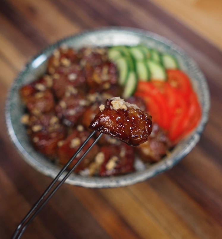 A glossy cube of shaking beef held by chopsticks, showcasing the rich glaze and crispy garlic topping against a backdrop of rice, cucumber, and tomato.