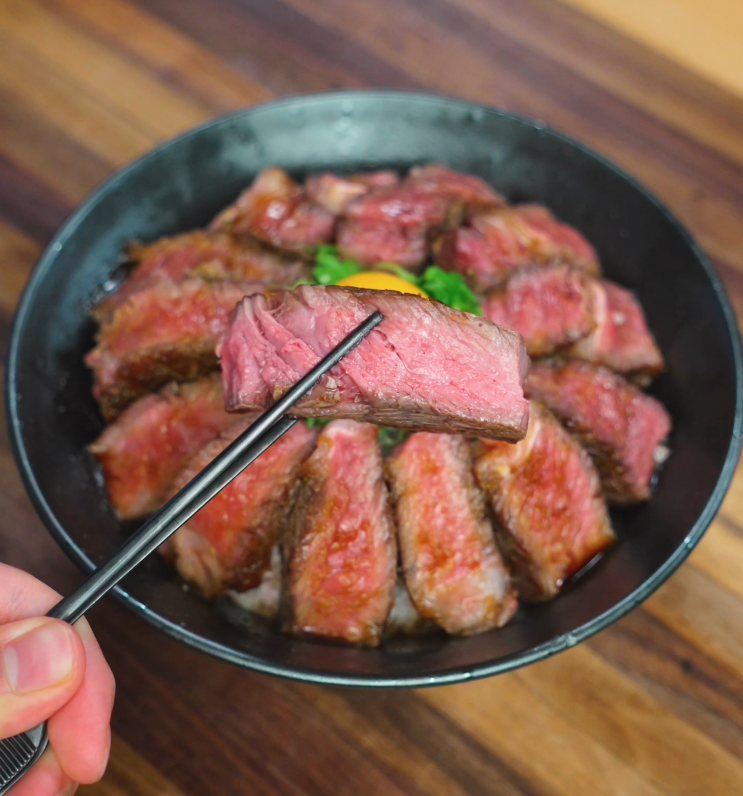 Chopsticks holding a juicy, medium-rare slice of steak above a rice bowl topped with egg yolk and scallions.
