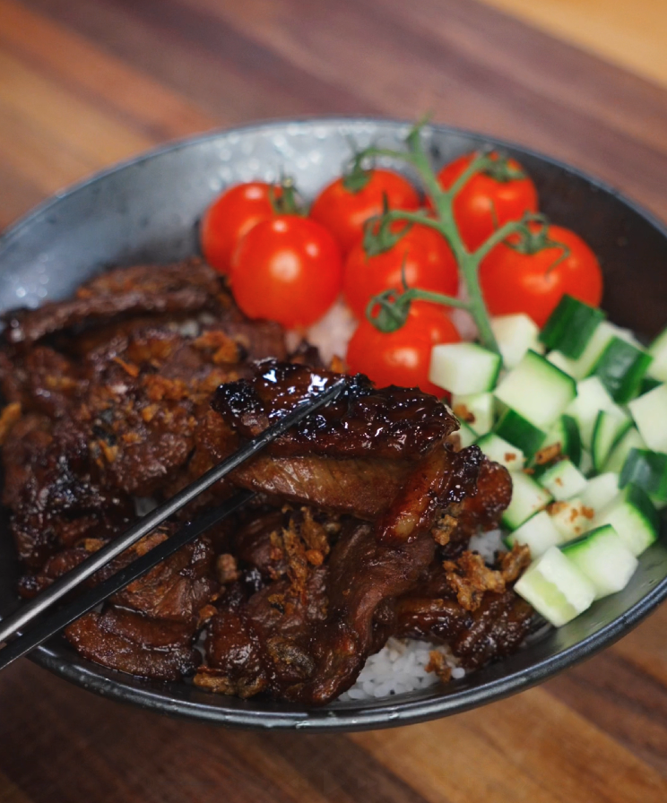 Lifting a glistening slice of beef tapa with chopsticks above the rice bowl showcasing its juicy, glazed texture.
