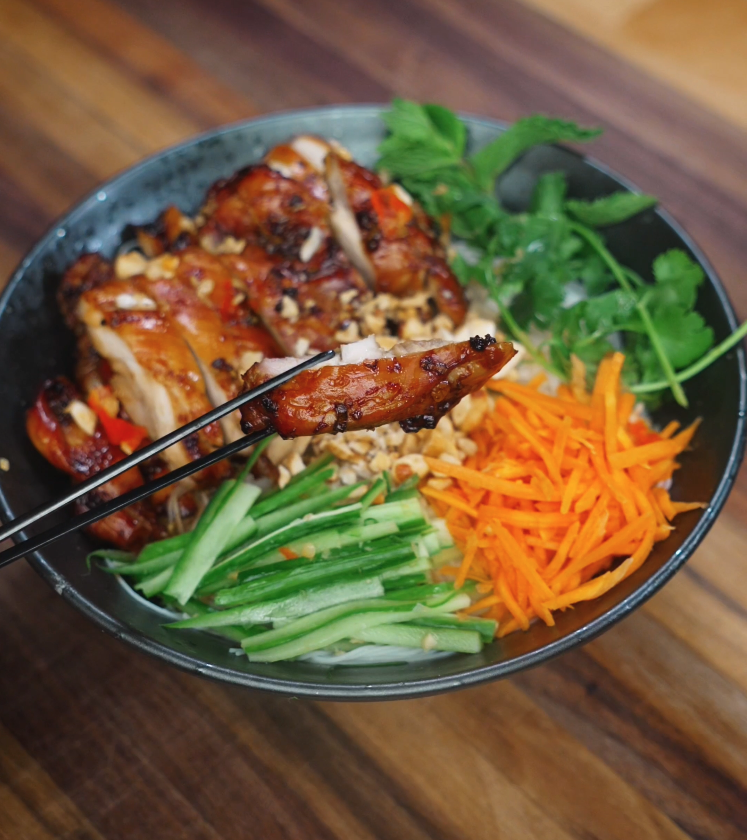 Closeup of a piece of grilled lemongrass chicken held with chopsticks above a colorful bowl of vermicelli, vegetables, and fresh herbs.
