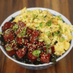 A vibrant close-up of the finished spam musubi rice bowl, topped with glossy caramelized spam cubes, soft scrambled eggs, scallions, and sesame seeds over a bed of white rice and seaweed.