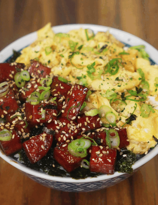 A vibrant close-up of the finished spam musubi rice bowl, topped with glossy caramelized spam cubes, soft scrambled eggs, scallions, and sesame seeds over a bed of white rice and seaweed.