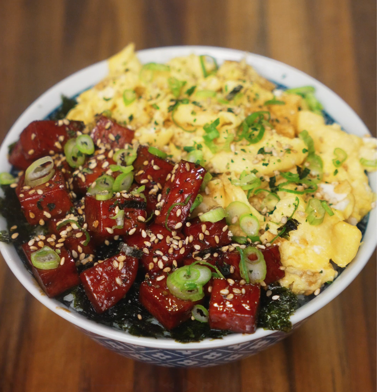 A vibrant close-up of the finished spam musubi rice bowl, topped with glossy caramelized spam cubes, soft scrambled eggs, scallions, and sesame seeds over a bed of white rice and seaweed.