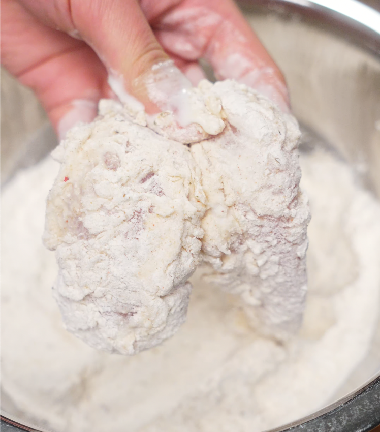 Hand pressing buttermilk-marinated chicken into a seasoned flour mixture, ensuring a thick coating for an ultra-crispy hot honey chicken crust.