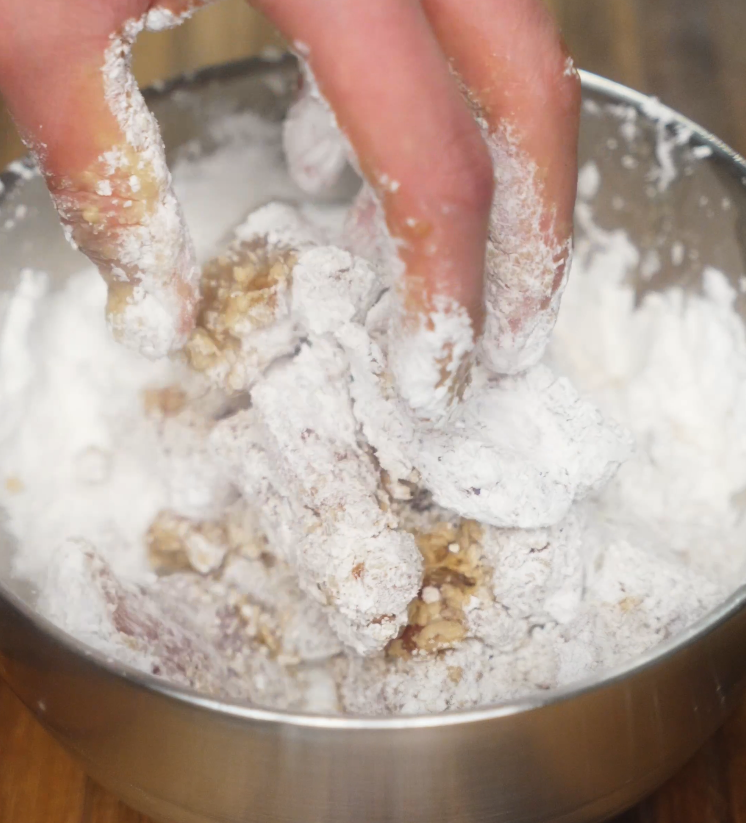 Hand mixing marinated chicken pieces in a metal bowl filled with sweet potato starch to create a light, crisp coating.