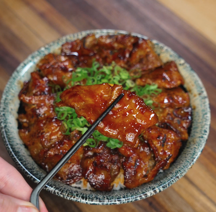 Close-up of glazed pork belly slice held with chopsticks over a bowl of rice topped with sticky spicy pork belly and chopped scallions, showcasing the glossy sauce and juicy texture.