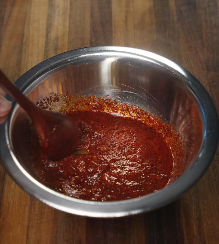 A thick, spicy kimchi jjigae sauce being stirred in a stainless steel mixing bowl. The deep red mixture contains gochugaru, garlic, and fermented ingredients, forming the flavor-packed foundation of the stew.