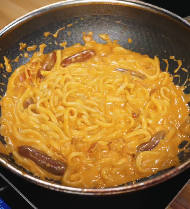 Freshly cooked rose noodles coated in a creamy pink tomato and cream sauce simmer in a nonstick pan, with pieces of sausage visible throughout.