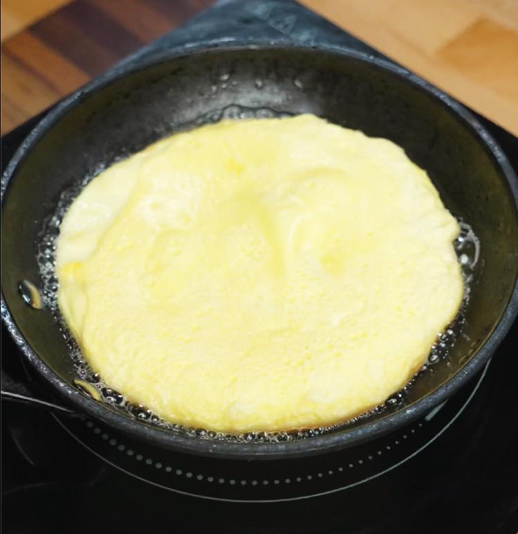 A thin egg crepe is being cooked in a skillet, forming a delicate layer that will later be sliced into strips for the somen salad topping.