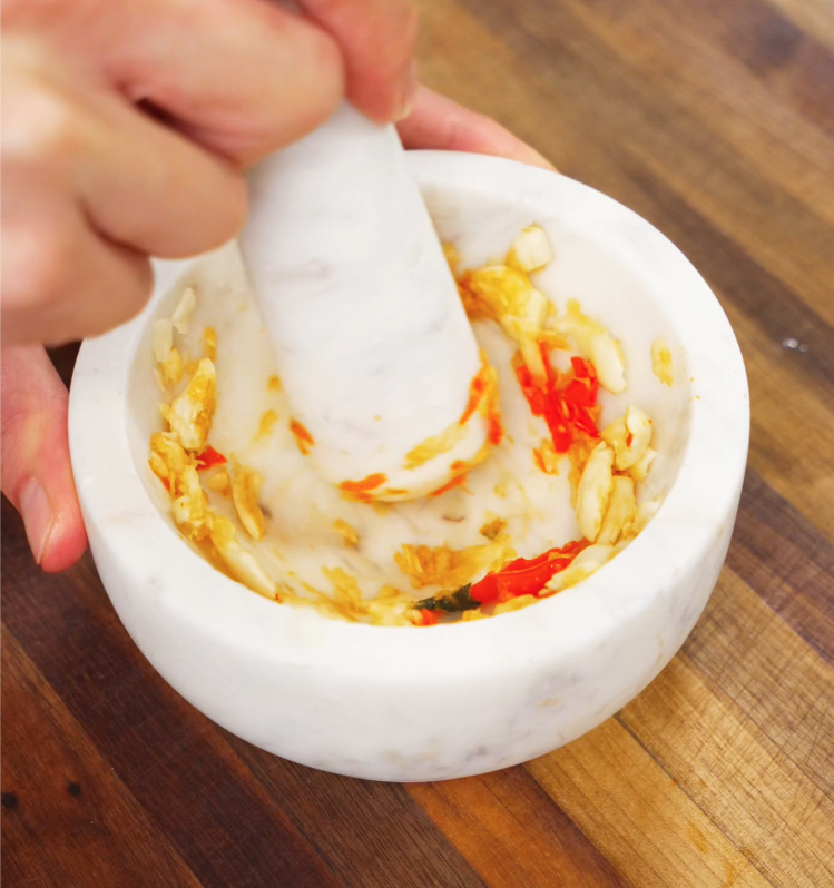 Fresh garlic cloves and red chilies are being crushed in a marble mortar and pestle to make a spicy Thai chili paste for authentic pad kra pao flavor.