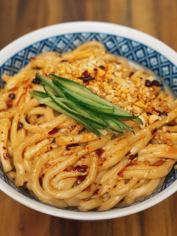 A close-up shot of creamy sesame noodles served in a patterned bowl, garnished with crushed peanuts, chili oil, and julienned cucumber for freshness.