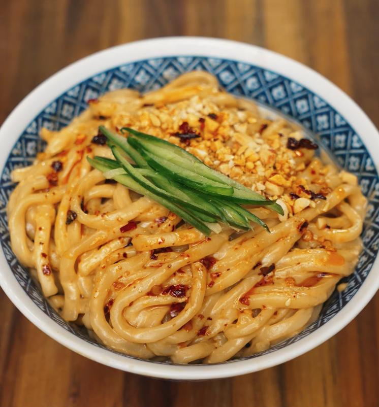 A close-up shot of creamy sesame noodles served in a patterned bowl, garnished with crushed peanuts, chili oil, and julienned cucumber for freshness.