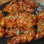 Close-up of crispy hot honey chicken tenders topped with sesame seeds and sliced green onions, served on a dark plate for a dramatic presentation.