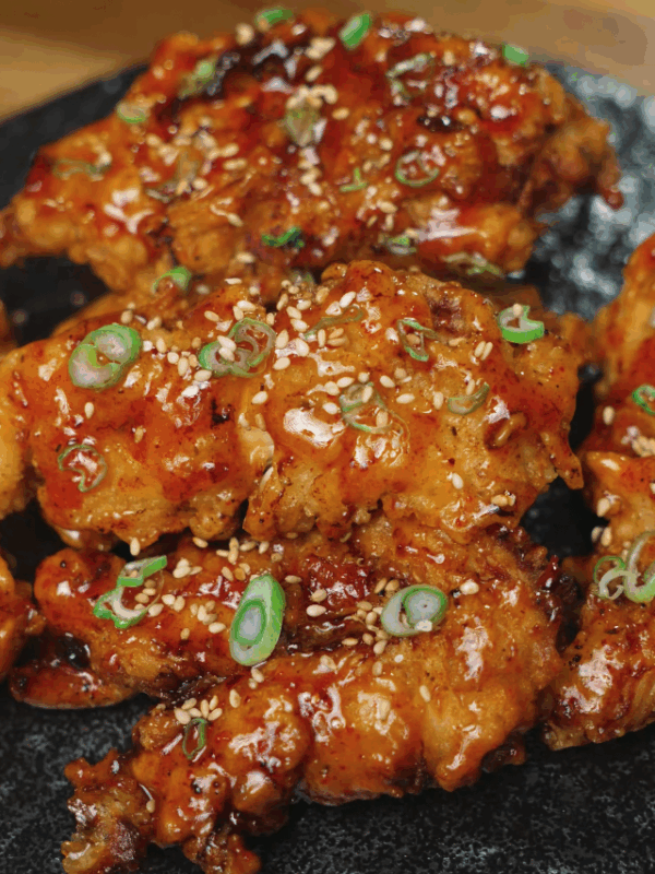 Close-up of crispy hot honey chicken tenders topped with sesame seeds and sliced green onions, served on a dark plate for a dramatic presentation.