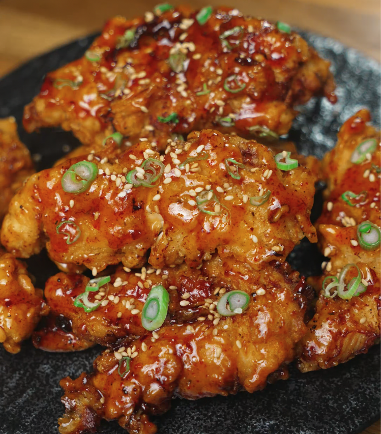 Close-up of crispy hot honey chicken tenders topped with sesame seeds and sliced green onions, served on a dark plate for a dramatic presentation.