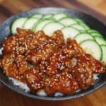 Crispy teriyaki chicken glazed in sauce, served over white rice with sliced cucumbers in black bowl