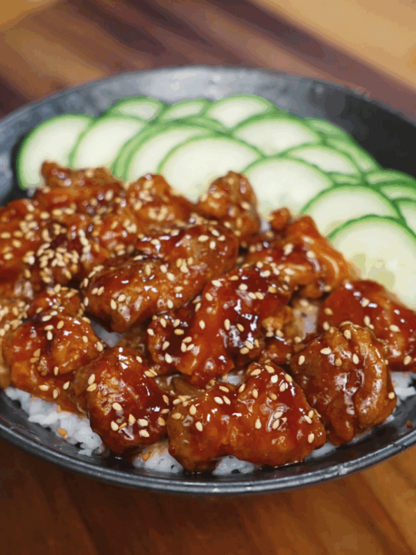 Crispy teriyaki chicken glazed in sauce, served over white rice with sliced cucumbers in black bowl