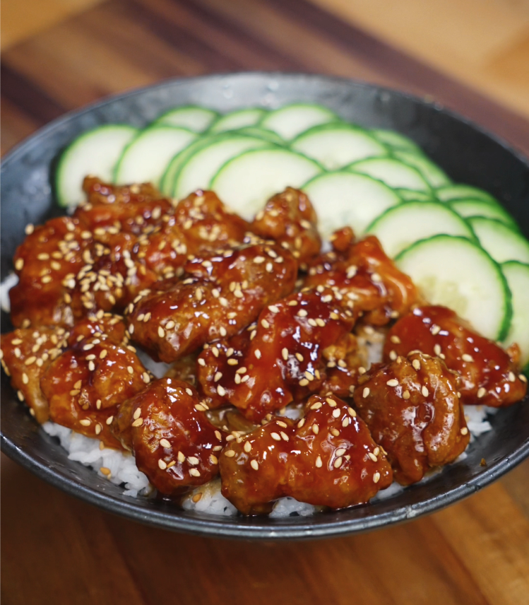 Crispy teriyaki chicken glazed in sauce, served over white rice with sliced cucumbers in black bowl