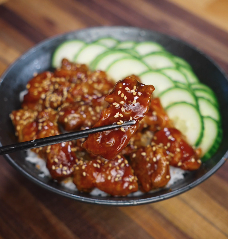 Close-up of crispy teriyaki chicken piece held with chopsticks over rice bowl with sliced cucumbers