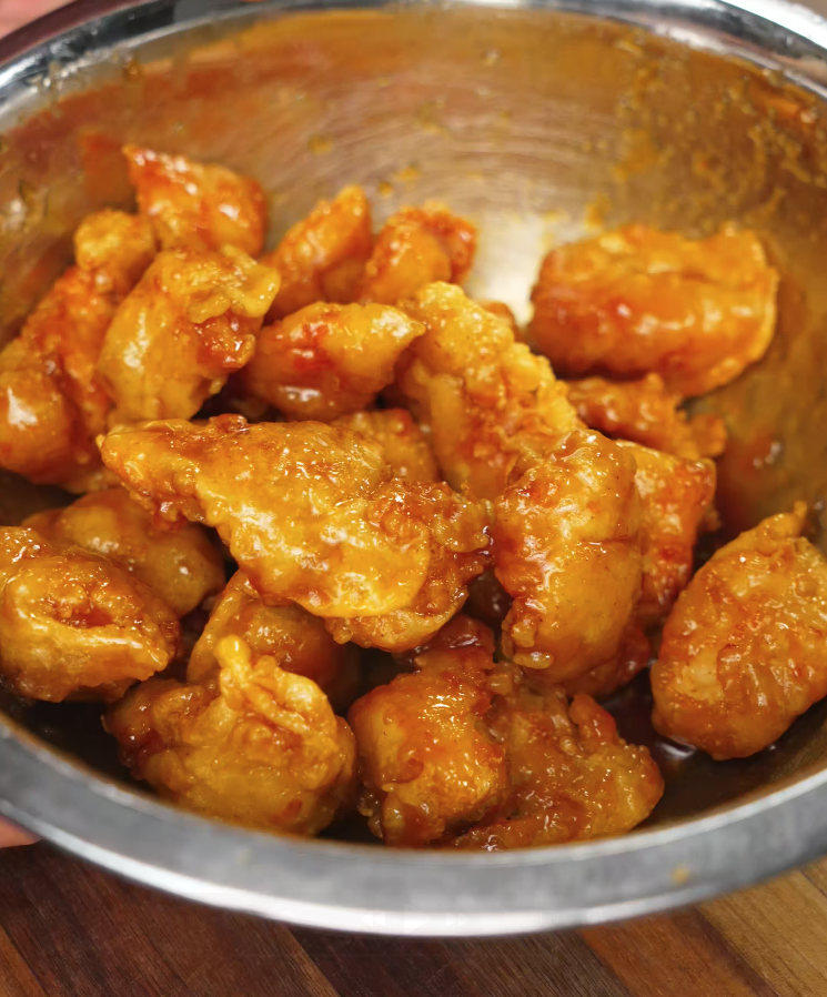 Golden fried chicken being coated in sticky sriracha honey butter sauce in a metal bowl