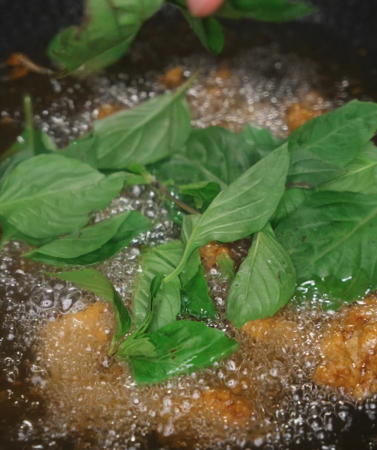 Fresh Thai basil leaves being dropped into hot oil, bubbling rapidly as they crisp up to add aroma and crunch.