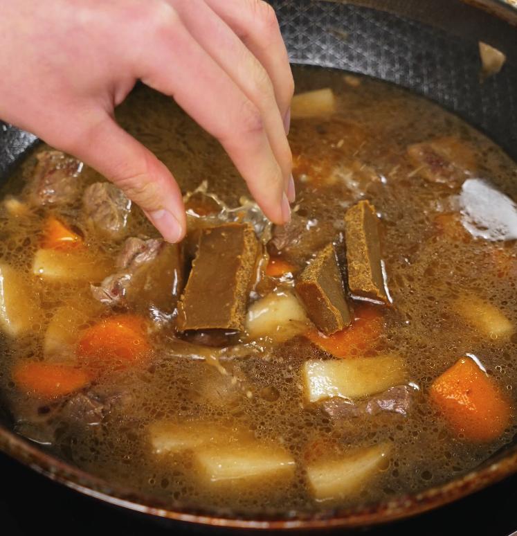 Curry roux blocks are being added into the simmering mixture of beef, potatoes, and carrots, where they will dissolve and thicken the Japanese curry.