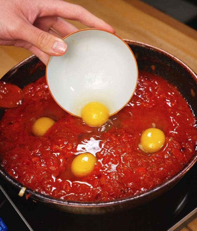Fresh eggs being gently cracked into the simmering tomato-chili base in a skillet, ready to poach in the rich, flavorful sauce.