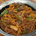 A bowl of glossy black pepper beef noodles with udon, tender beef slices, and green onions served in a rustic ceramic bowl.