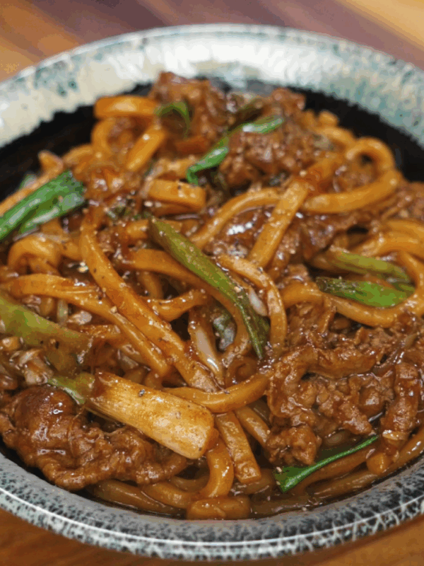 A bowl of glossy black pepper beef noodles with udon, tender beef slices, and green onions served in a rustic ceramic bowl.