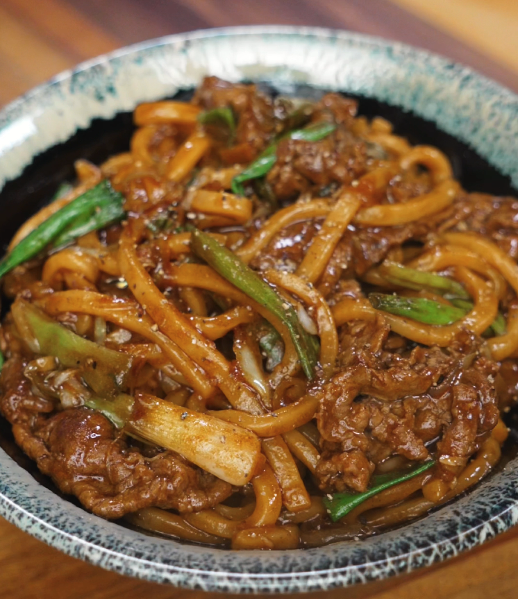 A bowl of glossy black pepper beef noodles with udon, tender beef slices, and green onions served in a rustic ceramic bowl.