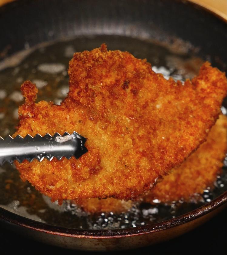 A golden brown breaded pork cutlet being deep fried in hot oil, showcasing the crispy texture and bubbling surface for the katsudon.
