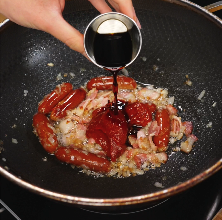A small metal cup of dark soy sauce is being poured into a pan filled with seared Chinese sausage, bacon, and softened onions to create a deep umami base for the rose noodle sauce.