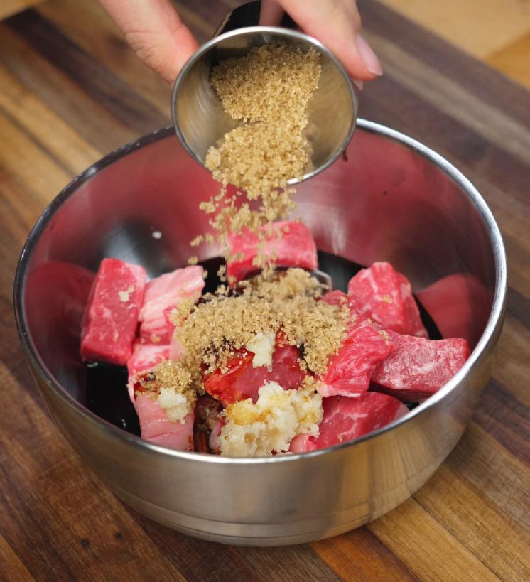 Brown sugar and minced garlic being added to a bowl of raw beef cubes to create a sweet and savory marinade, enhancing the depth of flavor for this shaking beef recipe.
