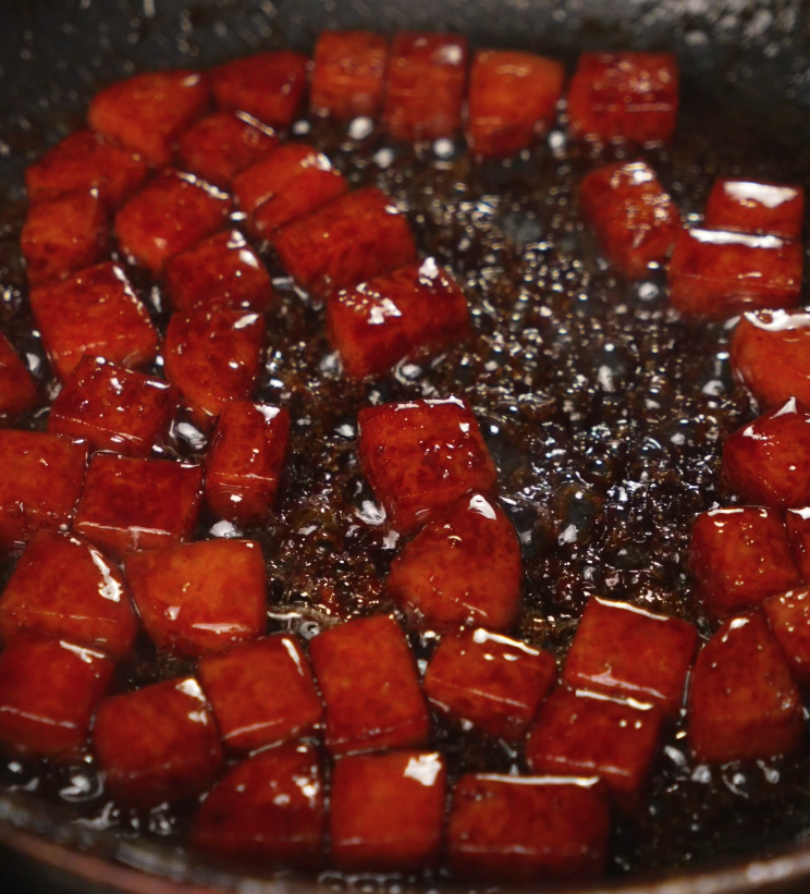 Spam cubes are tossed in a hot skillet with brown sugar and a rich savory sauce, beginning the caramelization process for the spam musubi rice bowl.