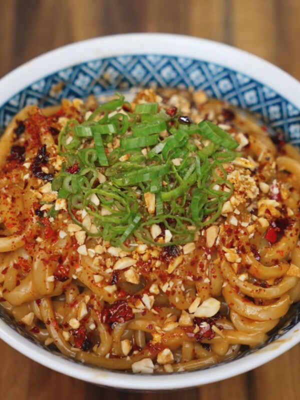 A close-up shot of spicy peanut butter noodles topped with chopped scallions, crushed peanuts, chili flakes, and chili oil, served in a patterned blue and white bowl on a wooden table.