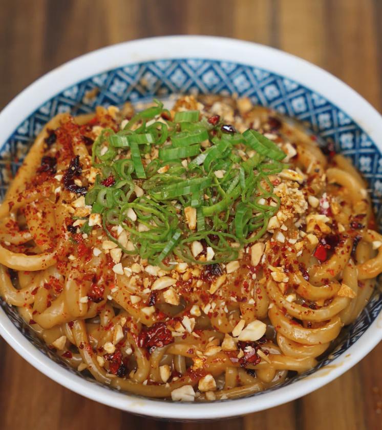 A close-up shot of spicy peanut butter noodles topped with chopped scallions, crushed peanuts, chili flakes, and chili oil, served in a patterned blue and white bowl on a wooden table.