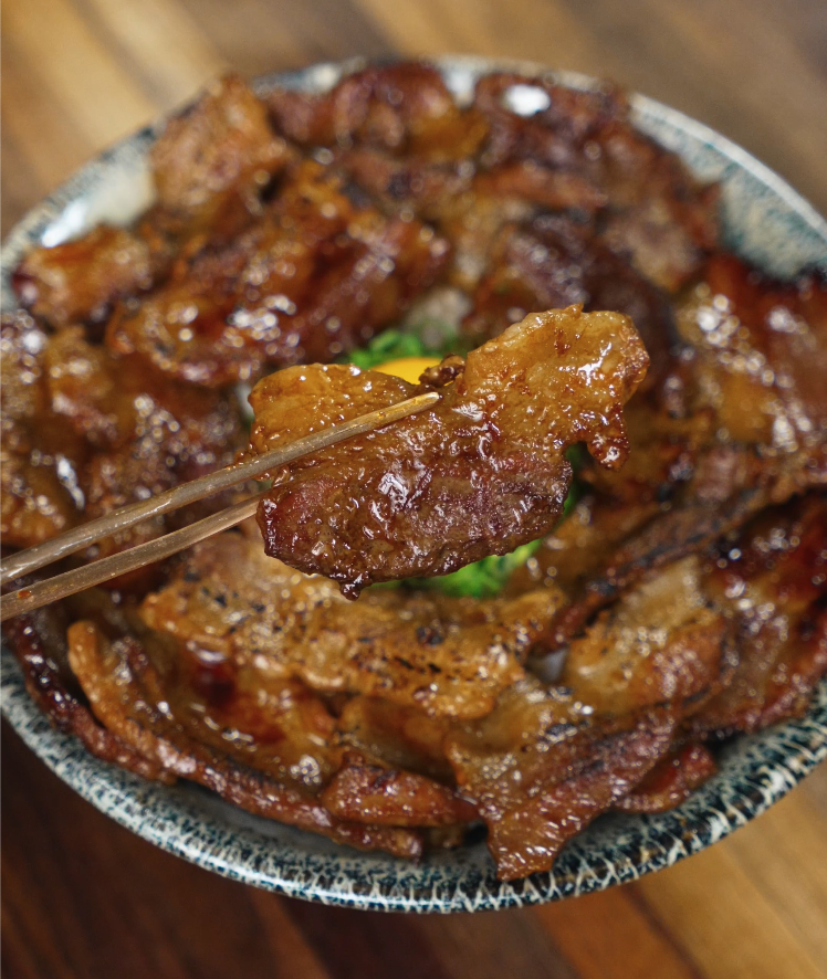 Close-up of glazed pork belly slice held with chopsticks over a bowl of rice topped with butadon pork belly and chopped scallions, showcasing the glossy sauce and juicy texture.
