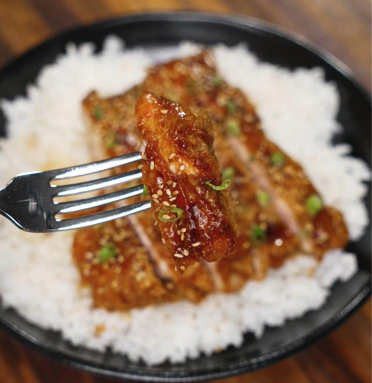 A fork holds up a juicy bite of honey lemon chicken topped with sesame seeds and scallions, showing the tender interior and glossy glaze.