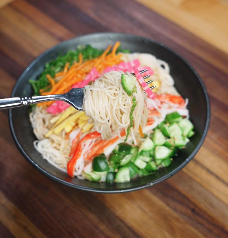 A close-up of a fork lifting chilled somen noodles with slices of cucumber and fish cake, showing the light and refreshing texture of the somen salad.