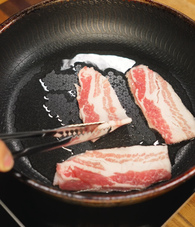Frying raw pork belly slices in a nonstick skillet with oil, showing the marbled layers of fat and meat beginning to render as they cook.