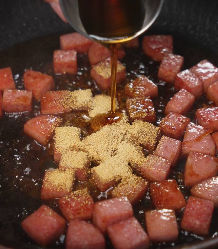 Spam cubes are tossed in a hot skillet with brown sugar and a rich savory sauce, beginning the caramelization process for the spam musubi rice bowl.