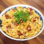 A high-resolution image of golden fried rice served in a patterned bowl, featuring diced lap cheong sausage, scrambled egg, and finely sliced green onions on top of the rice, sitting on a wooden surface.