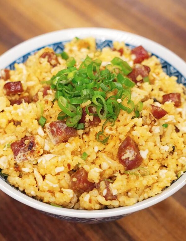 A high-resolution image of golden fried rice served in a patterned bowl, featuring diced lap cheong sausage, scrambled egg, and finely sliced green onions on top of the rice, sitting on a wooden surface.