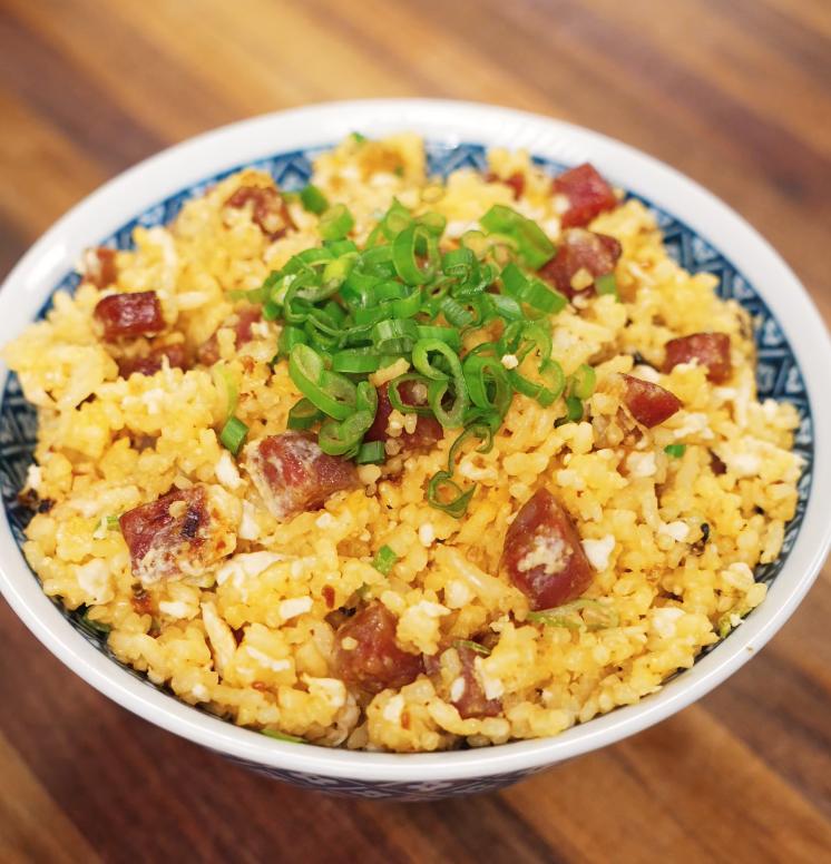 A high-resolution image of golden fried rice served in a patterned bowl, featuring diced lap cheong sausage, scrambled egg, and finely sliced green onions on top of the rice, sitting on a wooden surface.