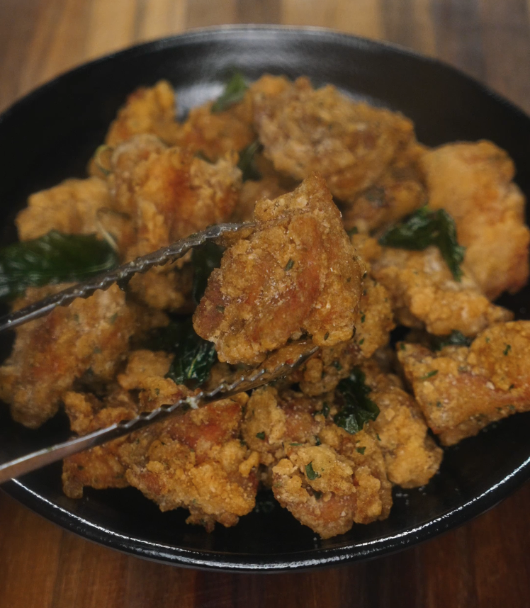 Close-up of crispy Taiwanese popcorn chicken held with tongs, showing texture and golden crust in front of plated dish.